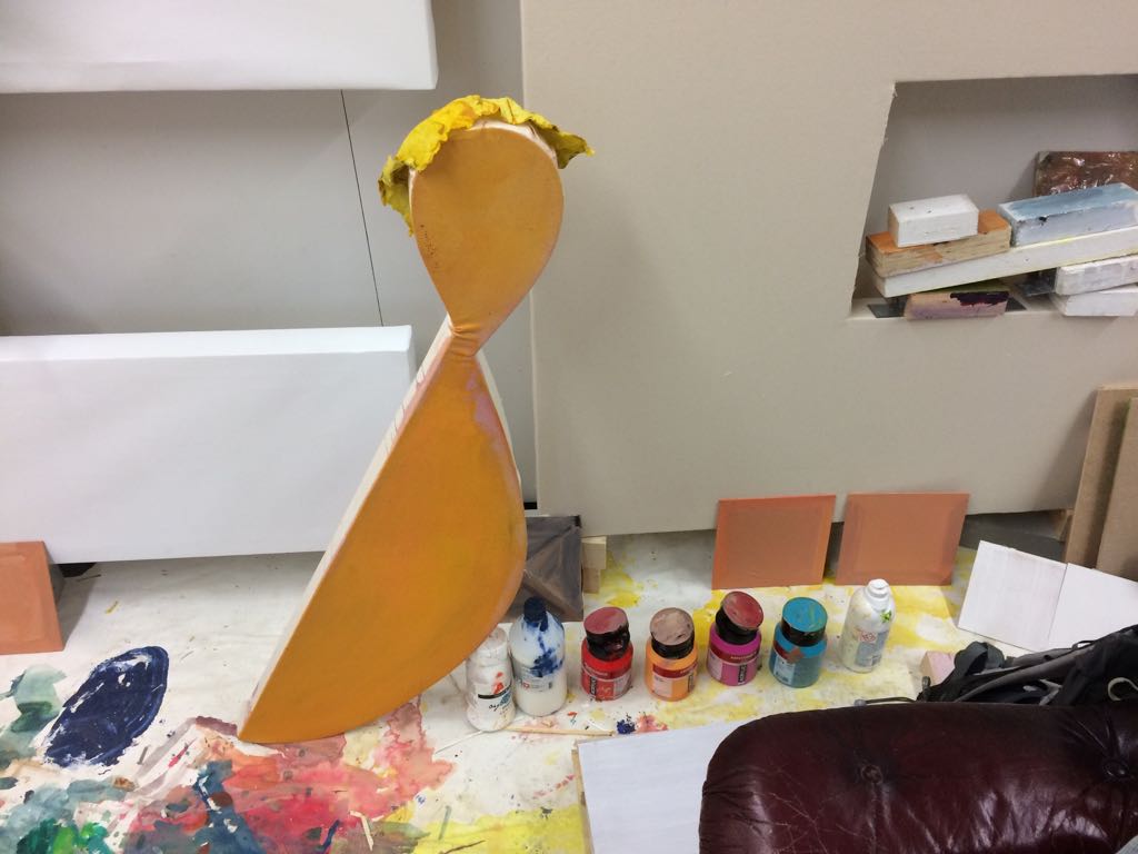 Kuva taiteilijan työhuoneelta. Lattialla on lukuisia maalipurkkeja, maalattuja kuvioita ja väritestien näköisiä läikkiä sekä geometrinen, keltainen litteä patsas.