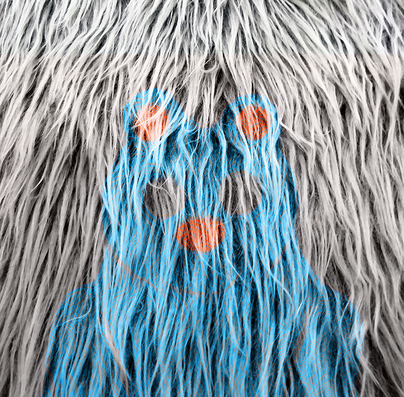 Lähikuva harmaasta turkista, jossa osa karvoisa on värjätty sinisellä ja punaisella. Värit muodostavat karvaan sinisen nallekarhun, jjolla on punaiset pallot korvina ja nenänä.