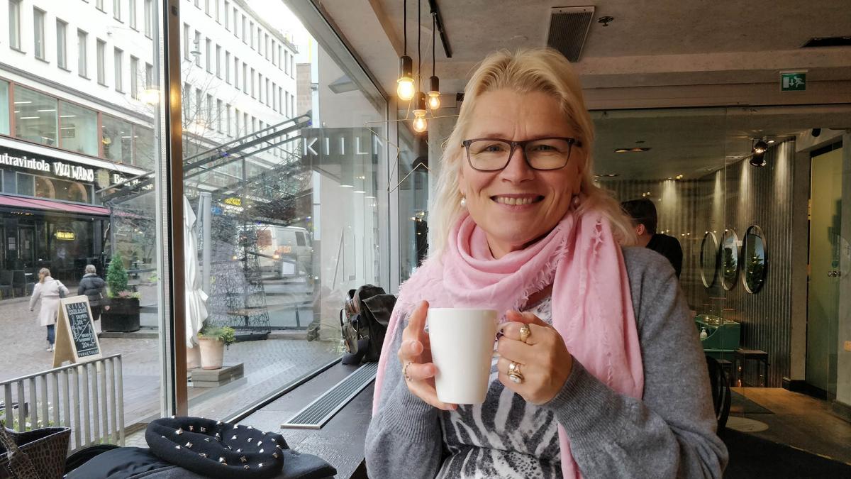 Laura Kiuru istuu kahvilassa kahvikuppi käsissään ja hymyilee kameralle. Taustalla on ikkuna, josta näkyy Helsingin keskustaa.