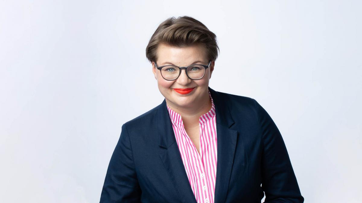 Lottaliina Pokkinen seisoo studiossa valleaa taustaa vasten ja hymyilee kameralle. Päällään hänellä on sininen jakku.
