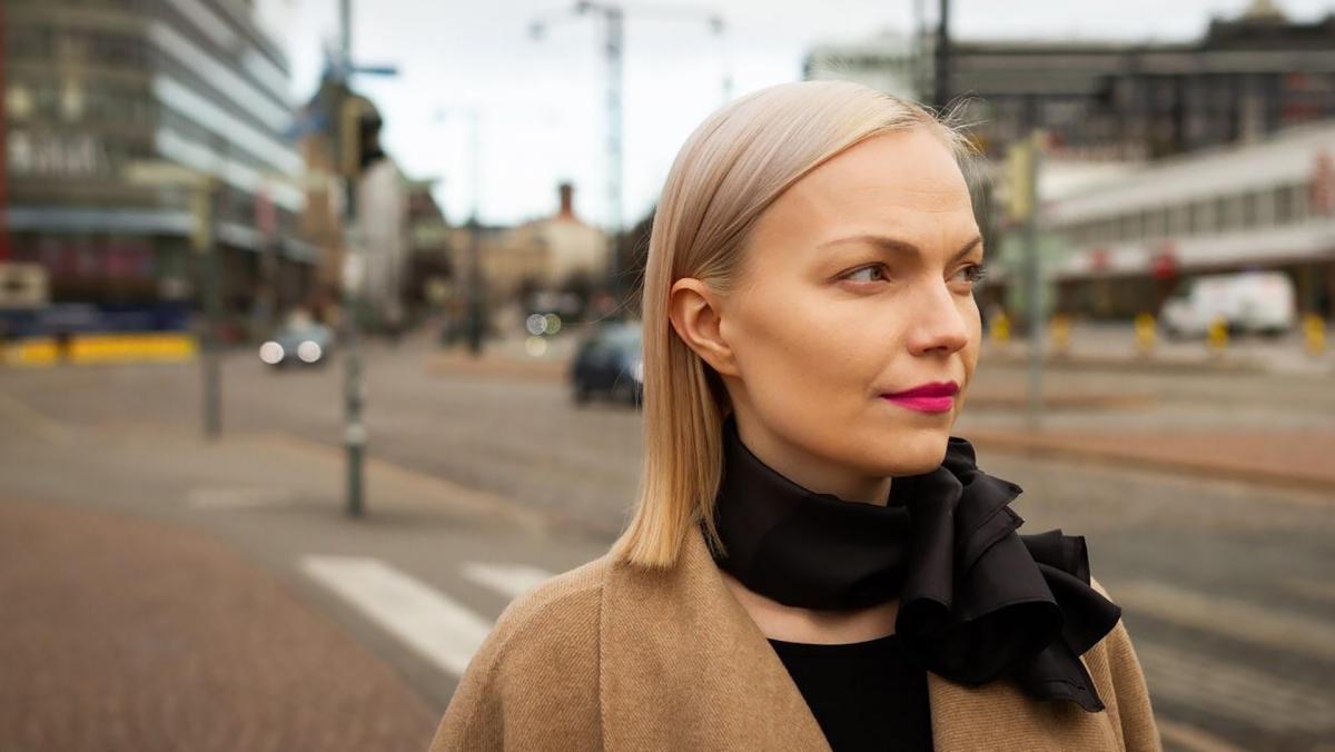 Minja Koskelan kasvokuva. Taustalla näkyy Helsingin keskustaa. Koskela katsoo kameran ohi oikealle neutraali ilme kasvoillaan.