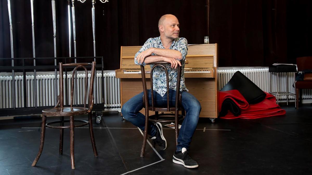 Jussi Tuurna istuu tuolilla teatterilavalla. Hän katsoo oikealle ja hymyilee. Hän istuu tuolissa väärinpäin, eli nojaa käsillään selkänojaan.