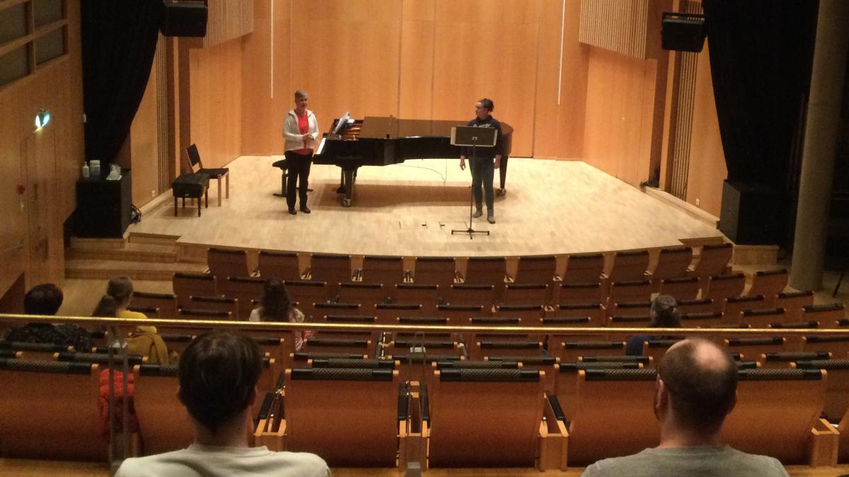 Laulunopettaja ja -oppilas Kaukametsän salin lavalla, katsomossa yleisöä