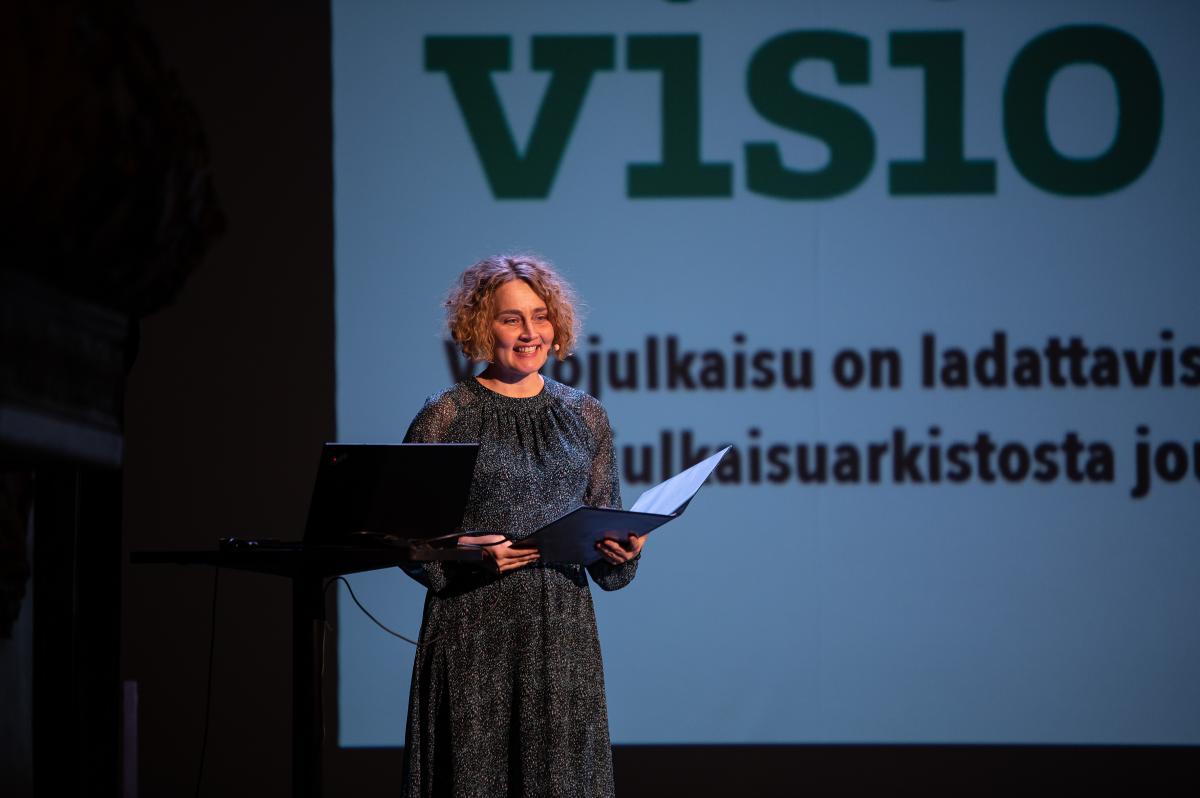 Keski-ikäinen hymyilevä nainen, jolla on kansio kädessään seisoo näyttämöllä. Taustalla lukee osa projisoidusta luentokalvosta. Siinä lukee osittaista tekstiä. Sanat visio ja julkaisu on lattavis ja julkaisuarkistosta erottuvat selvästi.