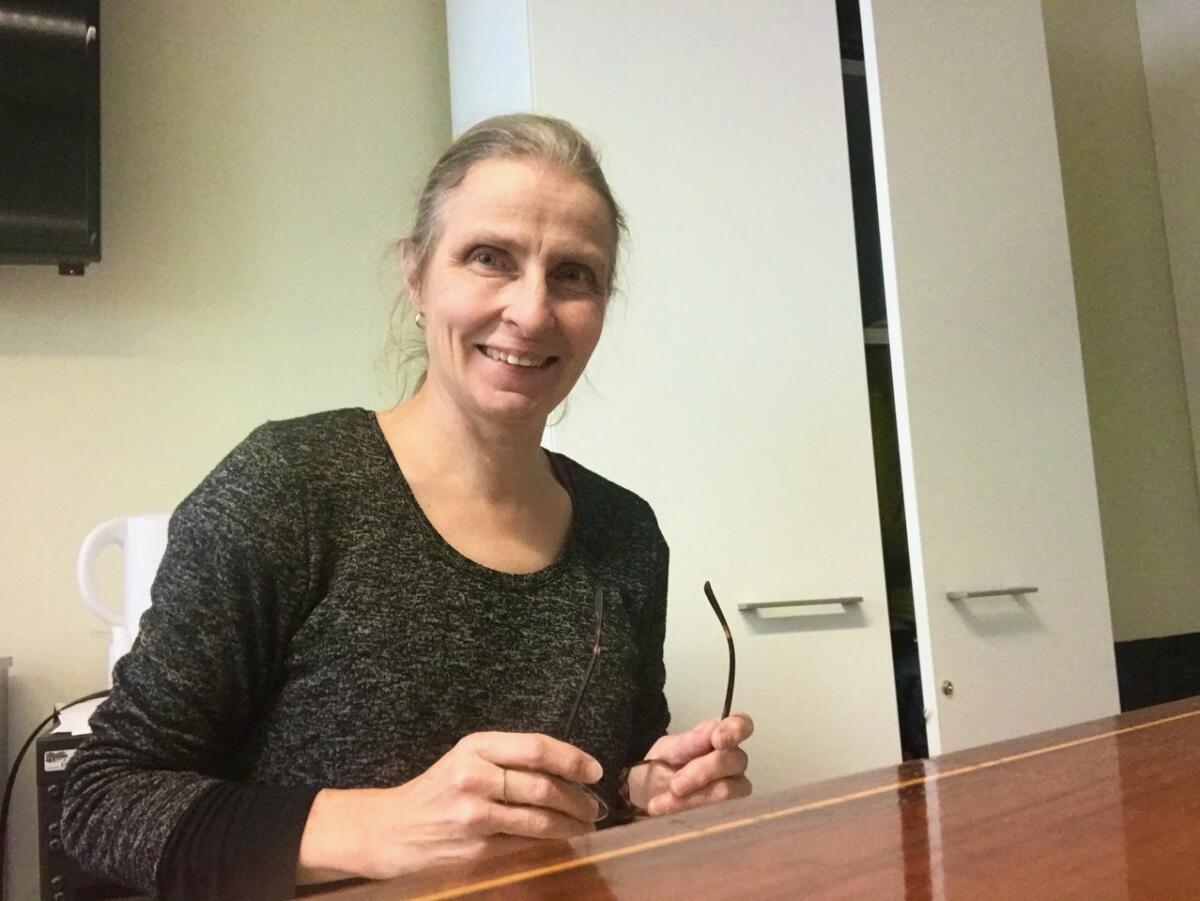 Sinikka istuu pöydän ääressä hymyillen pitäen kädessä silmälasejaan.