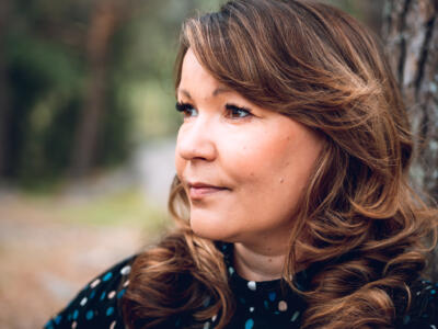 Vilma katsoo kuvasta sivulle mietteliäänä. Luontomaisema takana kuvassa on epätarkka.