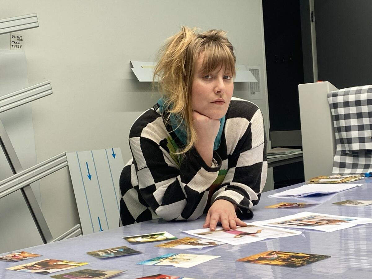 A white woman wearing a black and white print shirt leans on a table with printed photographs on it.