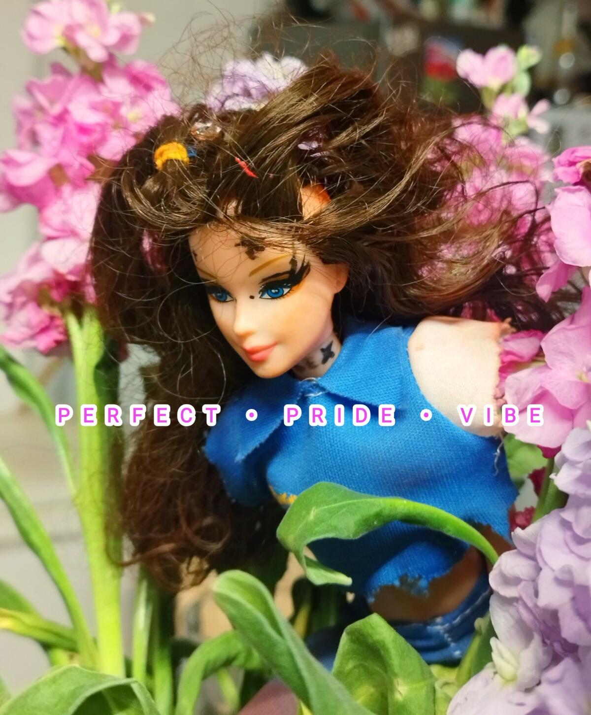 A close up shot of a barbie doll with flowers all around the doll. There are three texts in pink and with a white outline. They say "Perfect . Pride . Vibe".