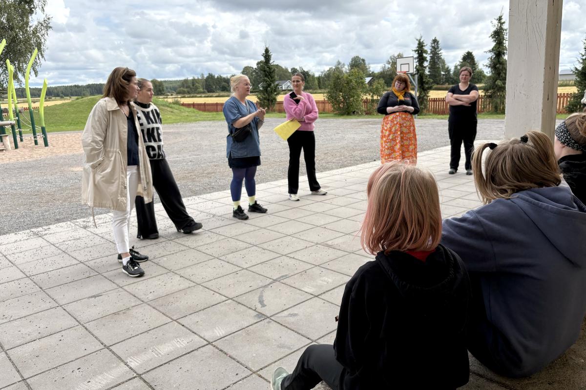 Ihmiset seisovat rivissä pihalla. Etualalla näkyy istuvia lapsia.
