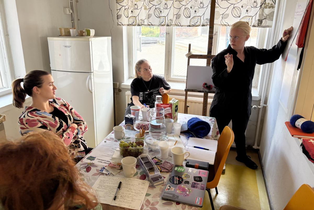 Kolme ihmistä istuu pöydän ääressä. Yksi seisoo ja kirjaa ylös asioita.