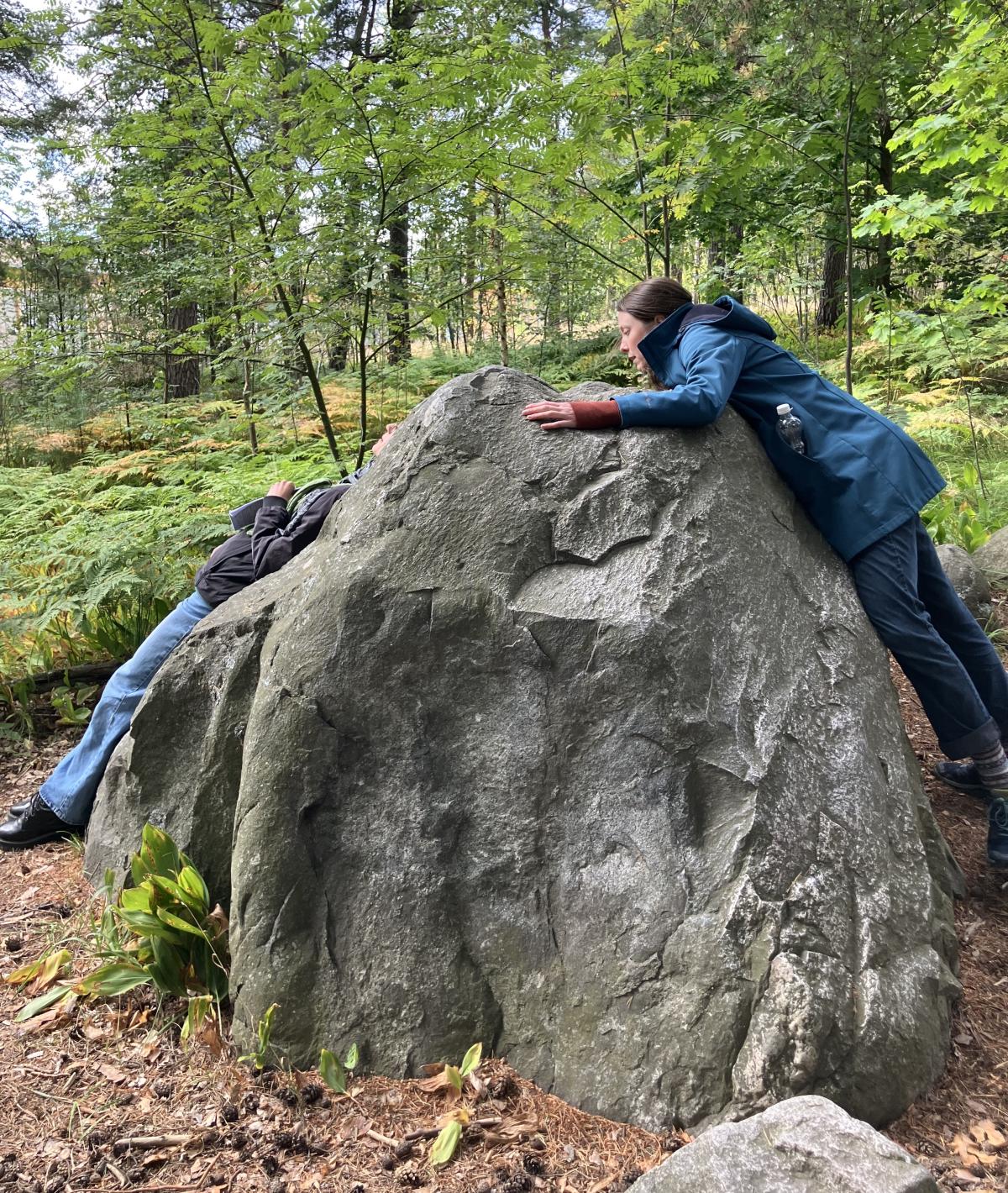 A person leaning with their front side on a big rock, antoher one leaning with their back.