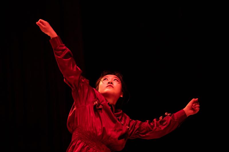 A dancer in red lighting spreading their arms and looking up.