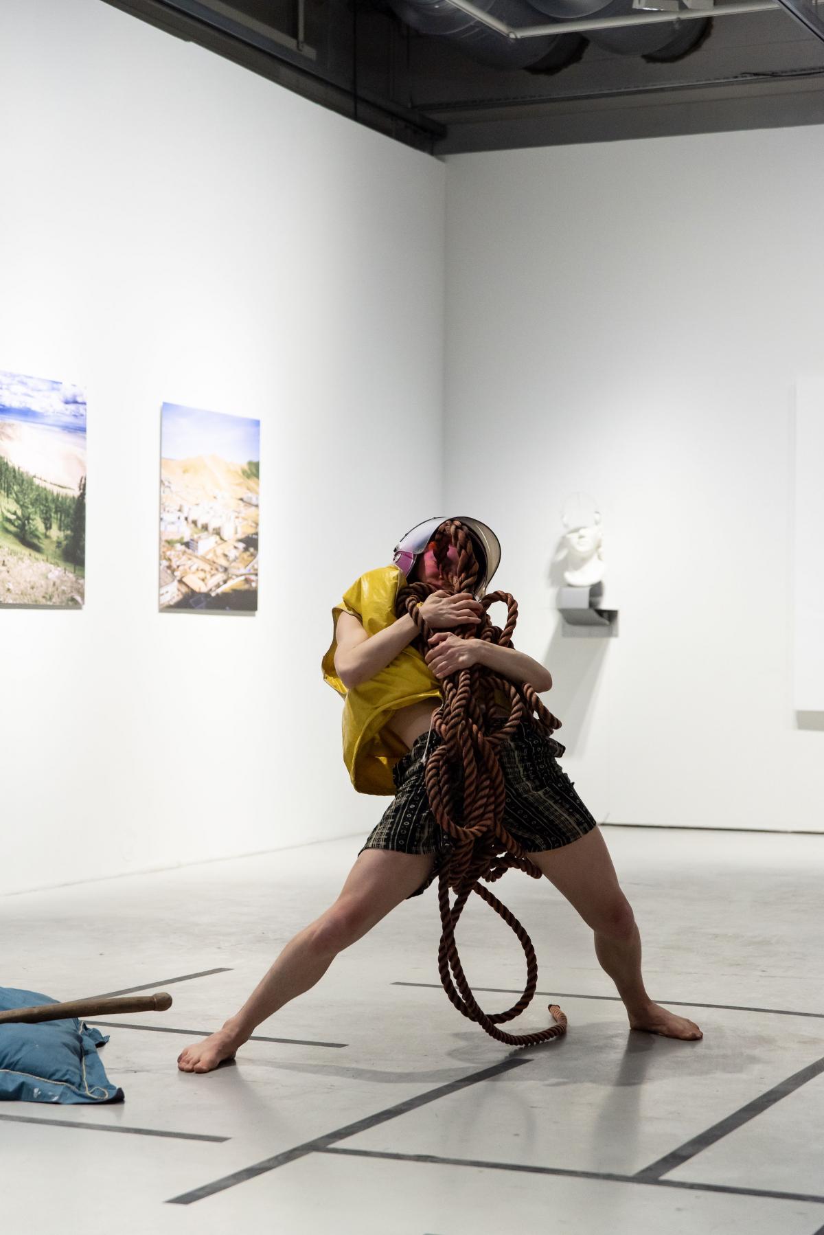 A person wearing a pink former police helmet and a yellow life jacket is holding a bundle of red sea rope close to their chest. The person has a wide stance and is leaning back in their posture. They are performing in a gallery with some artwork behind them. 