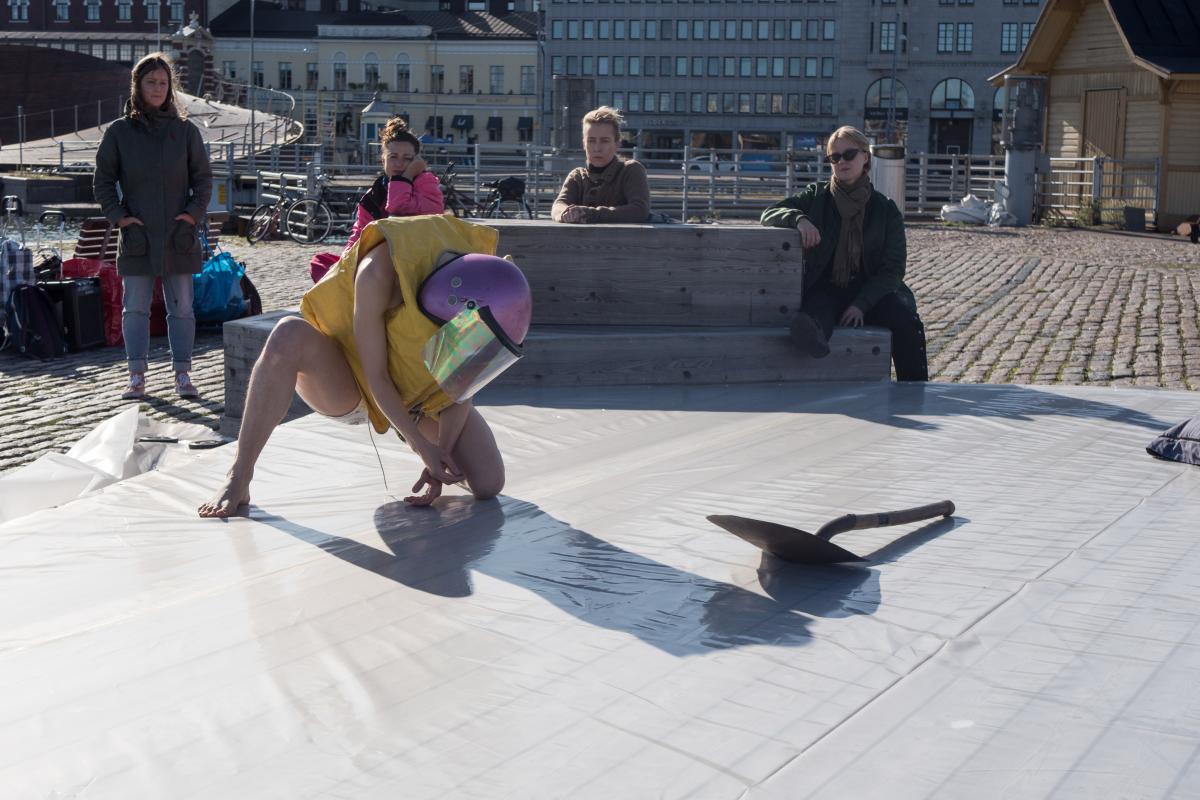 A person wearing a pink former police helmet and a yellow life jacket is squatting on the wooden stage outdoors in Kauppatori. There is a metal shovel next to the person. Several people are watching the performance.