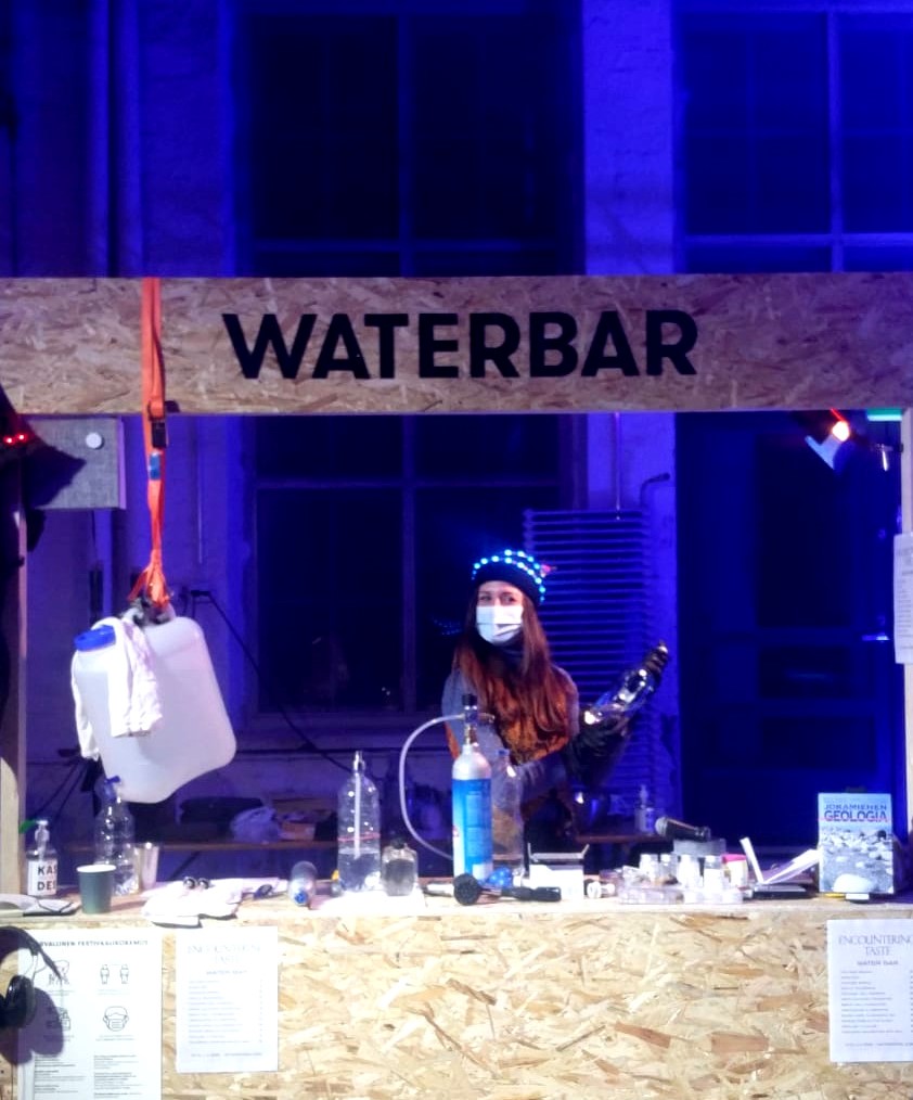 The picture shows the water bar set during ANTI festival Prize Party. At the water bar, you could taste waters from different areas of Finland and mineral waters synthesized using natural and artificial mineral samples. In the photo, artist Tea Andreoletti is behind the counter as she prepares sparkling water through a carbonization process.