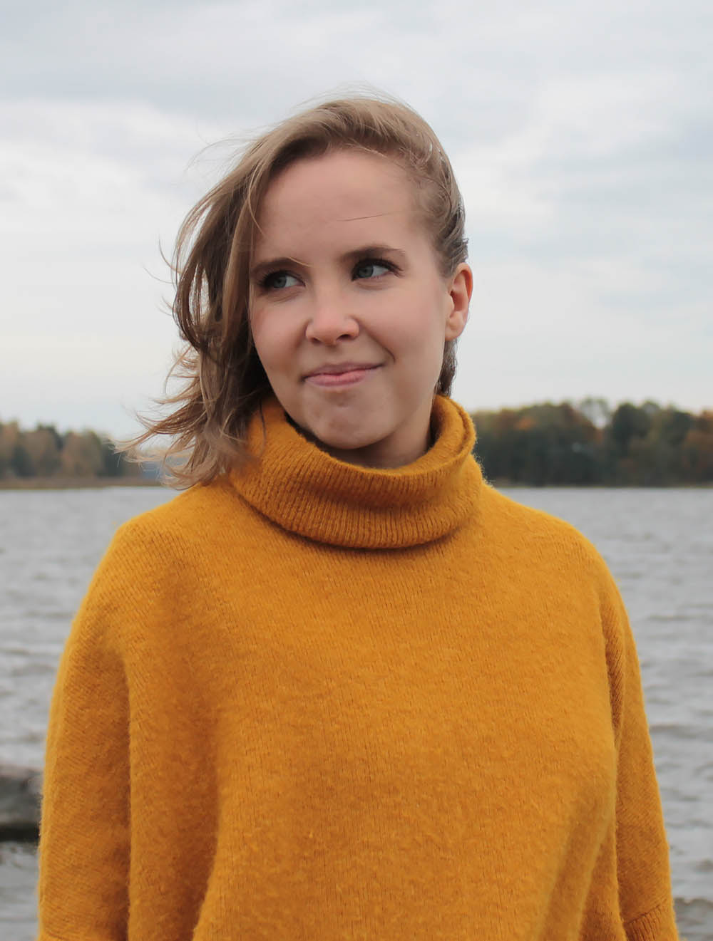 Johanna Rauma seisoo meren rannalla keltainen villapaita päällään ja katsoo vasemmalle hymyillen.