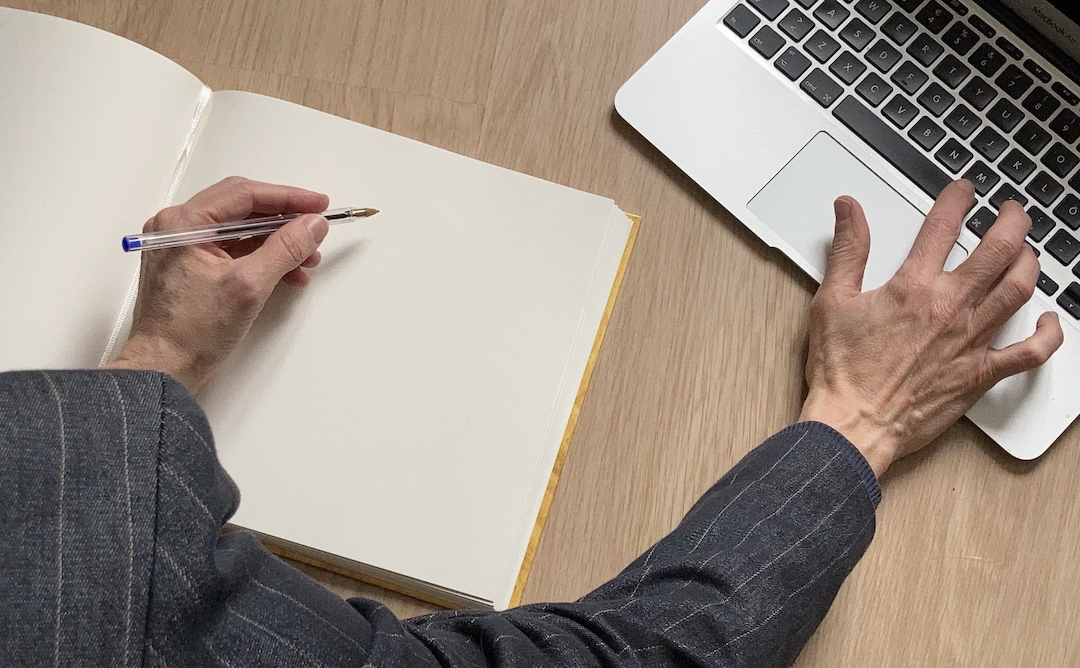 Left hand holding a pen on a notebook, right hand on the keyboard of a laptop.