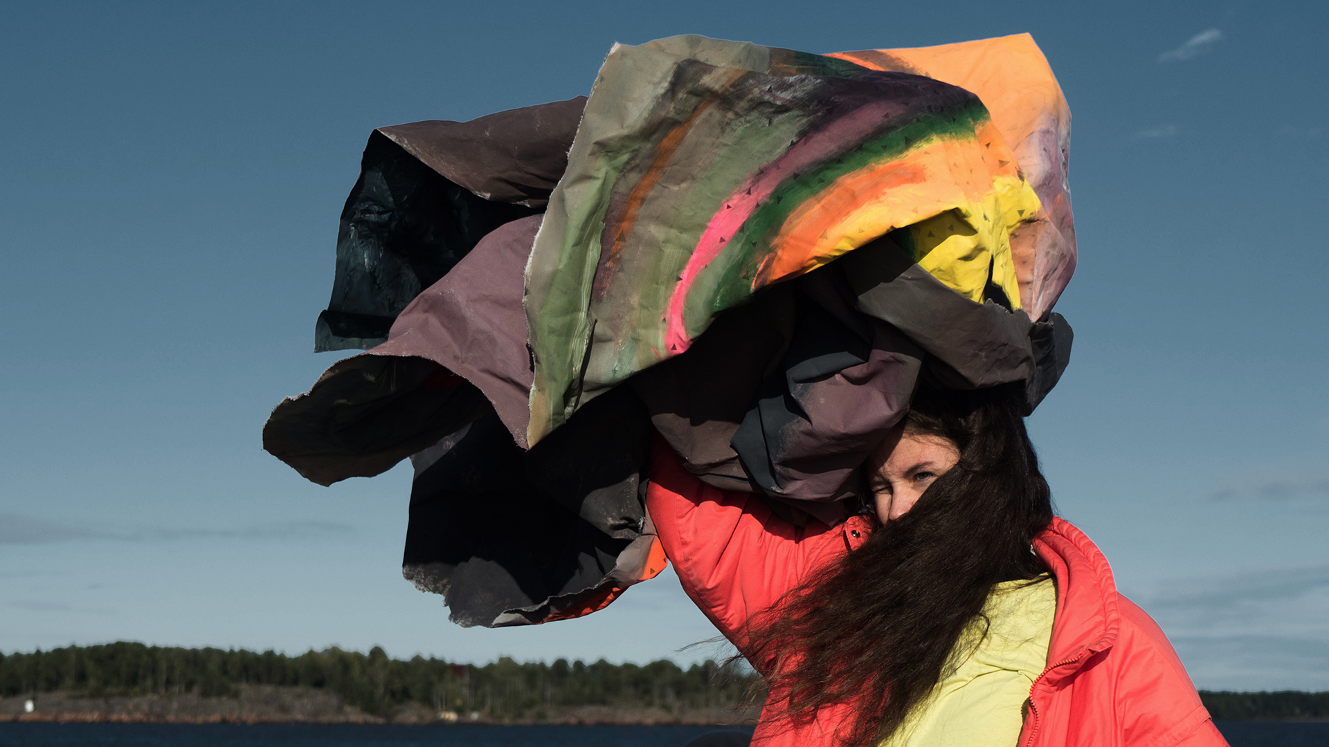 Long-haired performer outdoors holding a colourful fabric flapping in the wind above their head.