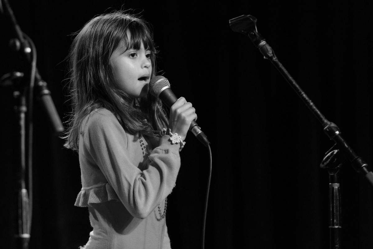 a black and white picture of a little girl singint to a microphone, dark background