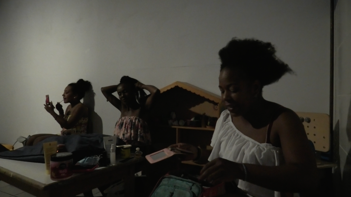 Three Afroperuvian women are putting on make-up and doing their hair. There are some cosmetic s on the table and a dollhouse at the background.