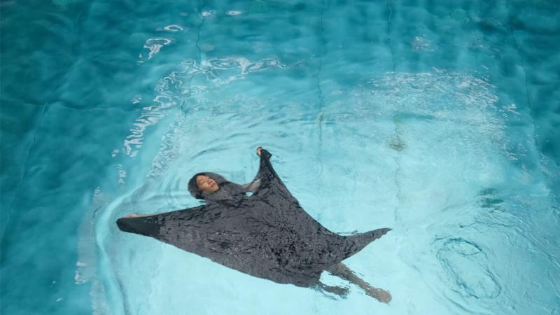 The artist is floating in water in a dark costume that is extended around him.