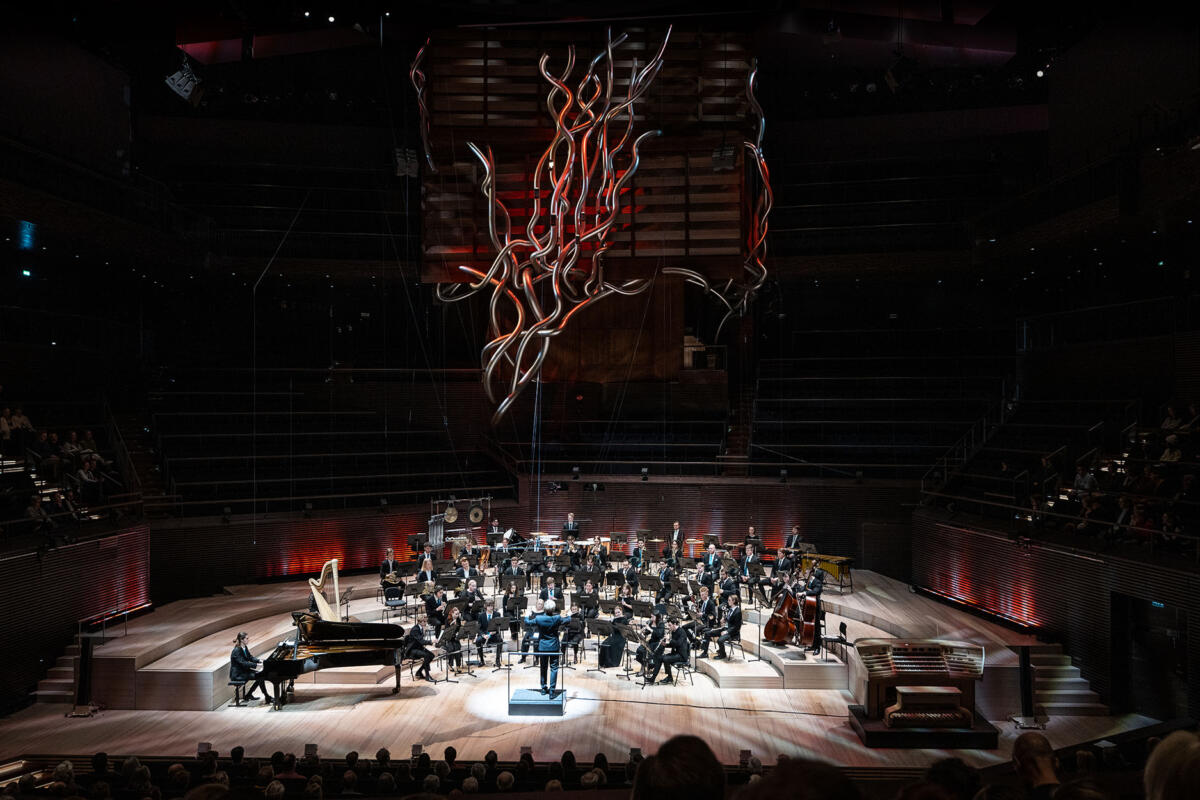 Kuva orkesterista kaukaa Musiikkitalossa. Urut hohtavat punaisina orkesterin yläpuolella pimeää katsomoa vasten.