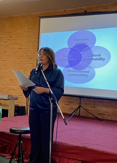 Vilma Timonen is standing behind the microphone stand talking. She has some papers on her hand. Behind on the screen there are four blue bubbles forming a fifth one in the centre. The bubbles have texts: Economic viability, cultural sustainability, environmental sustainability and social responsibility.