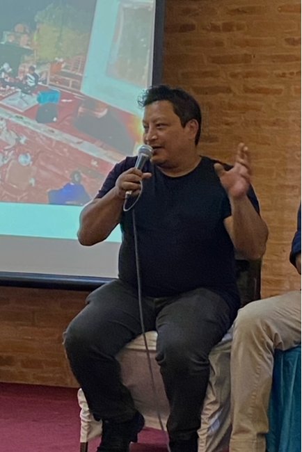 Pavit Maharjan sits on the stage with a part of a screen behind him. He is talking to a microphone on his hand.