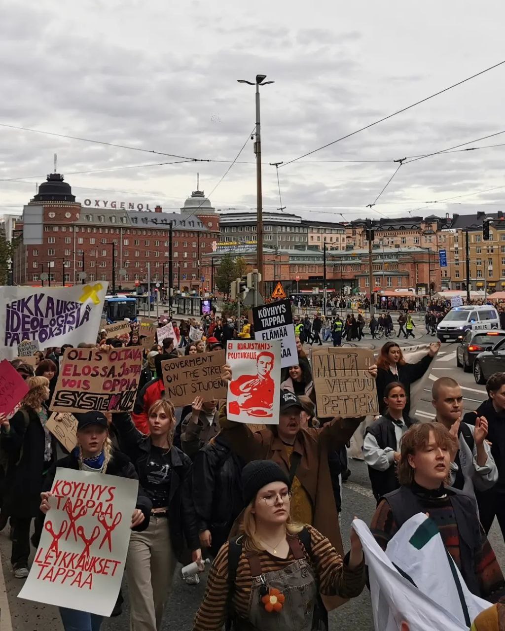 A large group of students is marching on Hämeentie. It's autumn. They are shouting and carrying signs saying: "The cuts are ideological decisions", "You should be alive, so that you can learn. System kills", "Government on the street, not students", "What the fuck Petteri", "No to the politics of austerity", and "Poverty kills, cuts kill"
